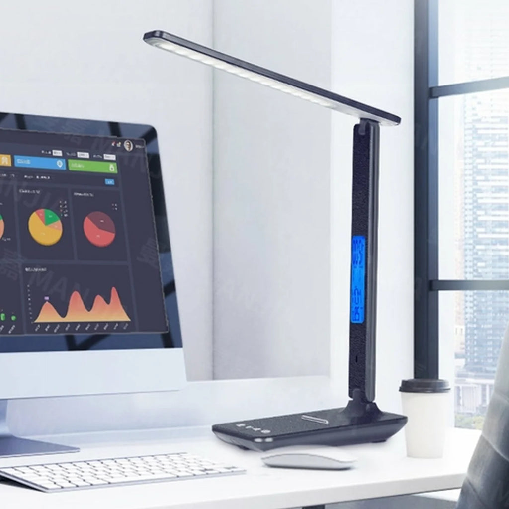 A black multi-functional desk lamp with built-in smartphone charging base, informational display screen, and illumination arm sitting on an office desk by a computer monitor, keyboard, and mouse.