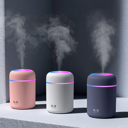 Three H2O portable humidifiers in pink, white, and dark gray, shown with mist emitting from their tops on a light gray surface against a dark gray background.