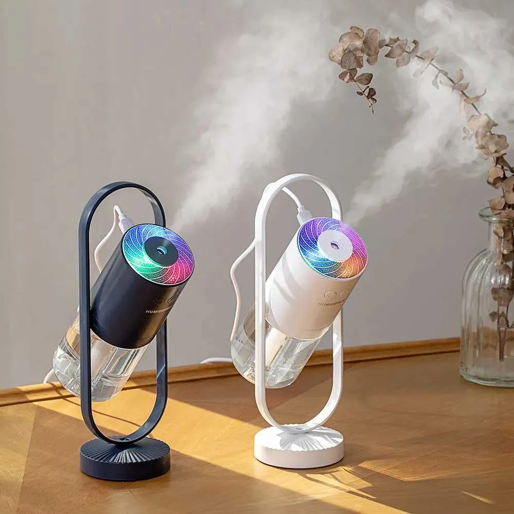 Two portable mist humidifiers with LED rainbow diffusers, one in black and one in white, shown on a wooden table with mist emitting and a vase of dried flowers in the background.