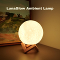 A glowing spherical moon lamp on a small wooden stand, placed on a tabletop in front of a framed landscape painting. The text 'LunaGlow Ambient Lamp' appears at the top.