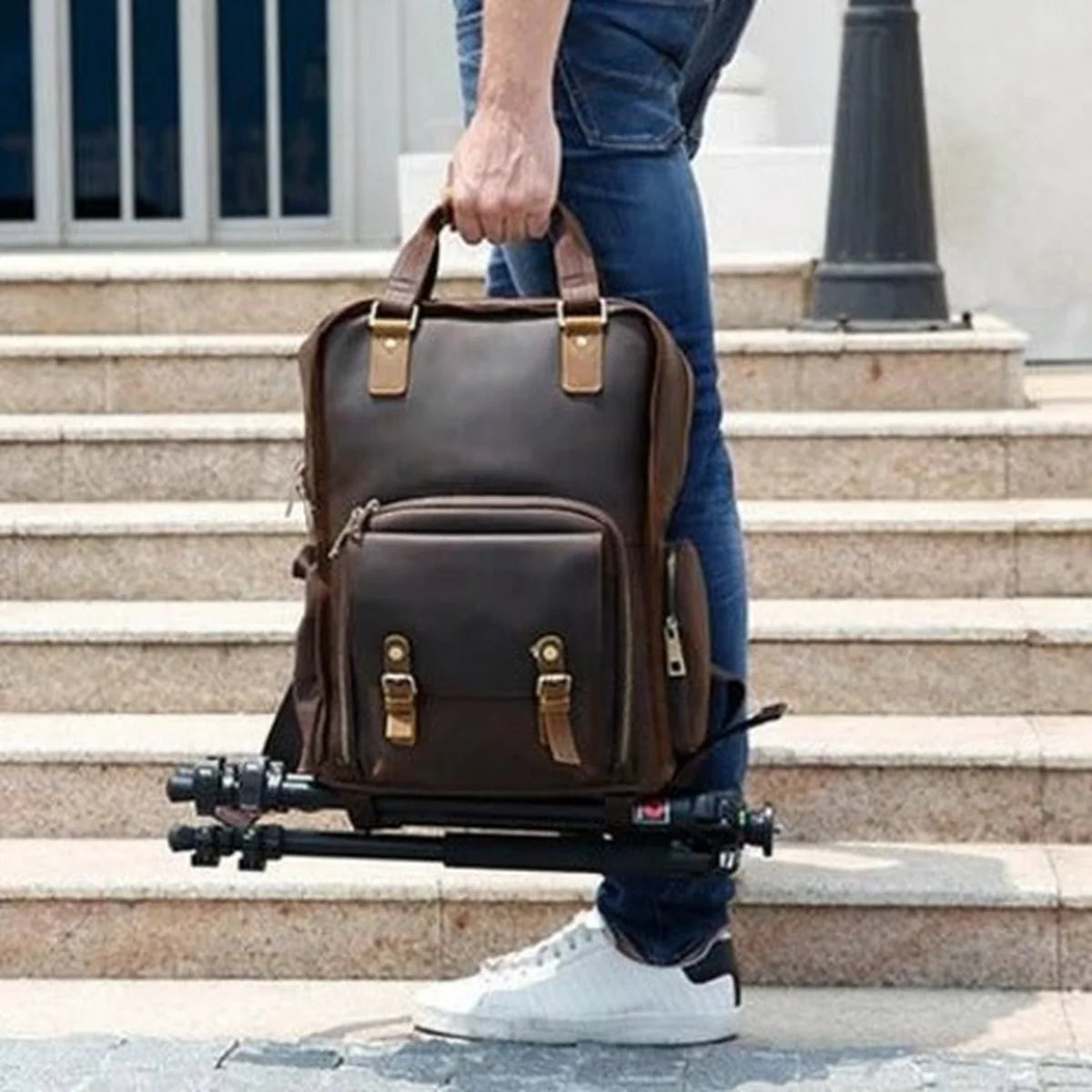 Person holding a large brown leather backpack with gold buckles on steps.
