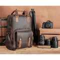 A brown leather camera backpack with tan leather accents stands on a wooden surface next to a tripod, camera, and lenses against a warm brown backdrop.