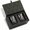 A pair of dark gray textured glass tumblers presented in an open black gift box with a gold leaf pattern on the interior lid and base.