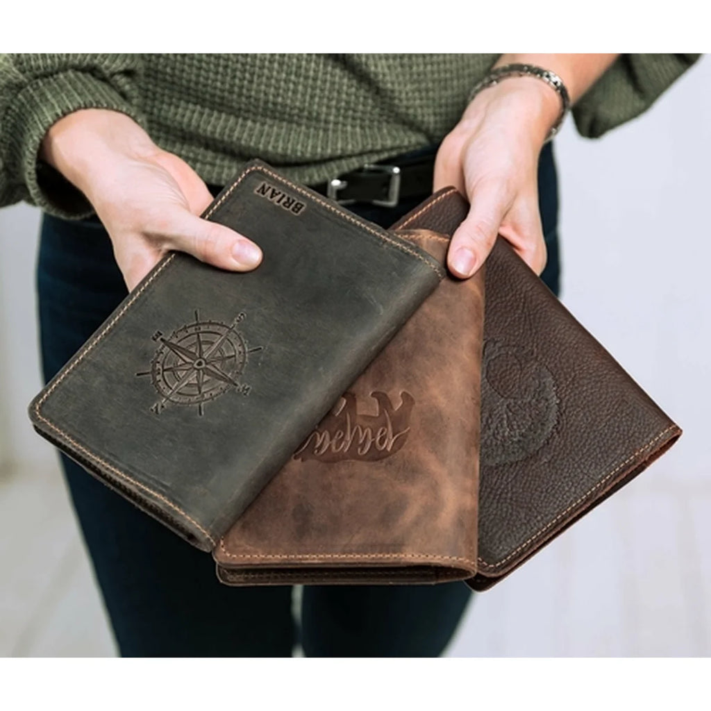 A person holds three leather passport covers in different brown shades, including a distressed dark brown one with an embossed compass design and a lighter brown one with an embossed crown.