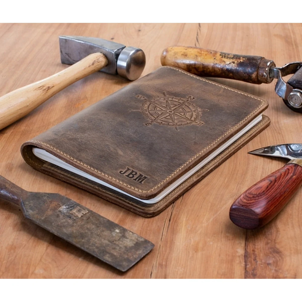 A brown leather notebook cover with an embossed compass rose and the initials 'JBM' on the front, styled on a wooden tabletop with tools including a hammer, chisel, and knife.