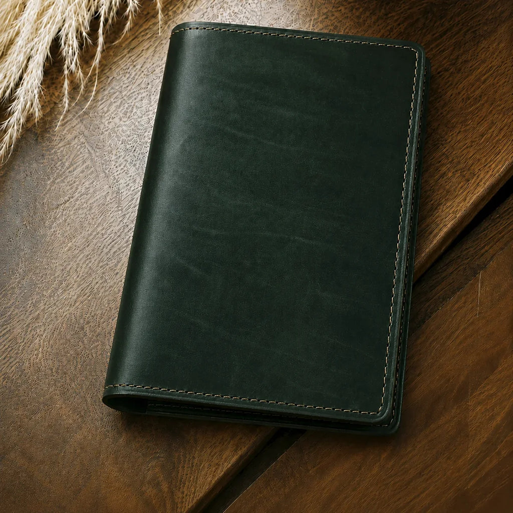 A dark forest green leather notebook or planner cover with light tan stitching, resting on a warm brown wooden surface with dried grass in the corner.