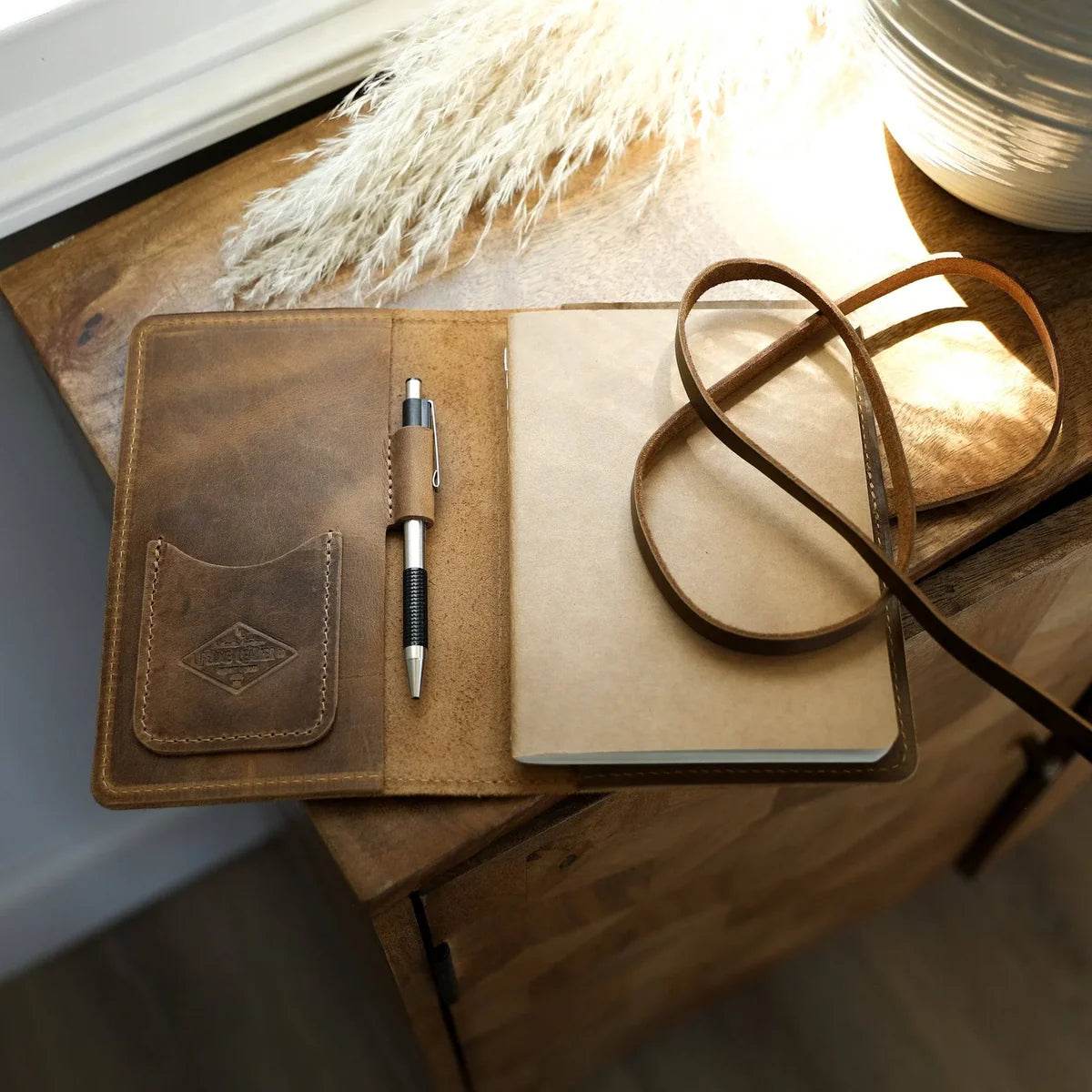 A brown leather notebook cover laid open on a wooden table, holding a beige notebook, a pen, and a long leather strap. A warm light from a nearby lamp casts a glow over the scene.