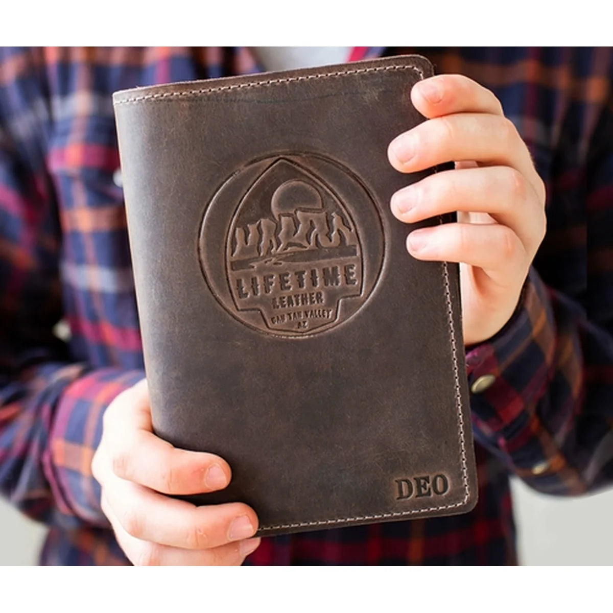 A person holding a dark brown leather wallet or notebook cover with an embossed circular logo reading 'LIFETIME LEATHER' and the initials 'DEO' in the bottom right corner. The person is wearing a red and blue plaid flannel shirt.