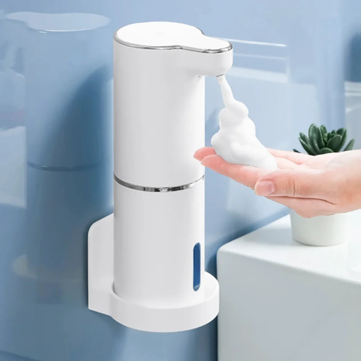 A white wall-mounted automatic soap dispenser with a touchless sensor and a hand receiving foam under a clear spout, set against a light blue bathroom wall.